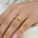 A close-up of a hand wearing a delicate gold ring with three small circular stones, including one green stone and one clear stone, set against a soft light background.