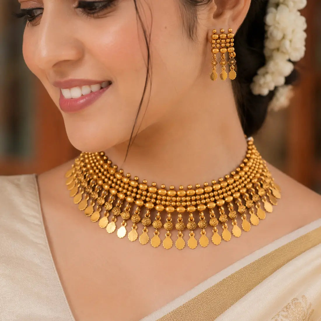 A close-up of a woman wearing a gold-toned traditional necklace with dangling disc charms and matching dangling earrings, paired with a cream-colored sari.