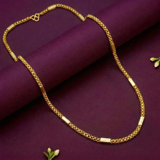 A gold chain necklace with rectangular bar links, draped elegantly over a deep purple rolled fabric backdrop with small white flower accents.