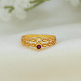 A gold ring with a textured, twisted band design, featuring a top setting with a clear round gemstone and a bottom setting with a round purple gemstone, displayed on a soft white fabric background.